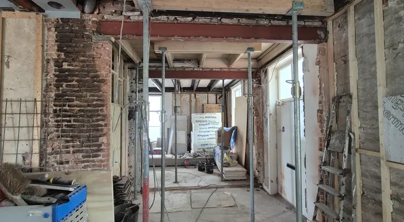 Voor: gestripte ruimte met stalen balken, tijdelijke bouwstempels, open wanden en isolatieplaten, gezien in de lengte van de woning